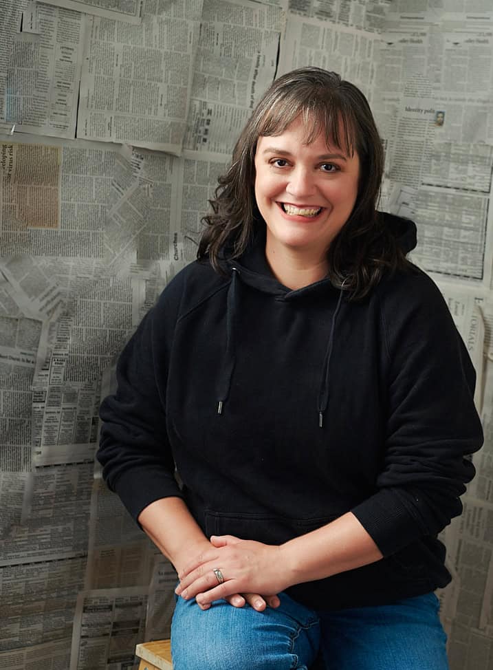 Jody E. Skinner is a caucasian woman with silver-streaked dark hair, and she is sitting in front of a wall of newsprint wearing a black hoodie and comfortable jeans.